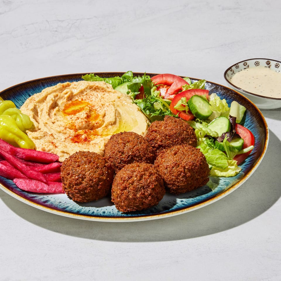 A blue plate with falafel, hummus, salad, pickled turnips, and peppers, with a side of sauce.