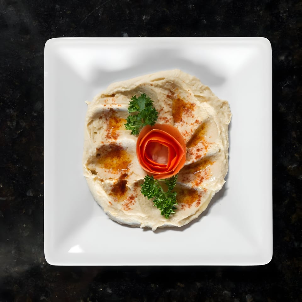 Creamy hummus on a white square plate, garnished with parsley, paprika, and a carrot flower.