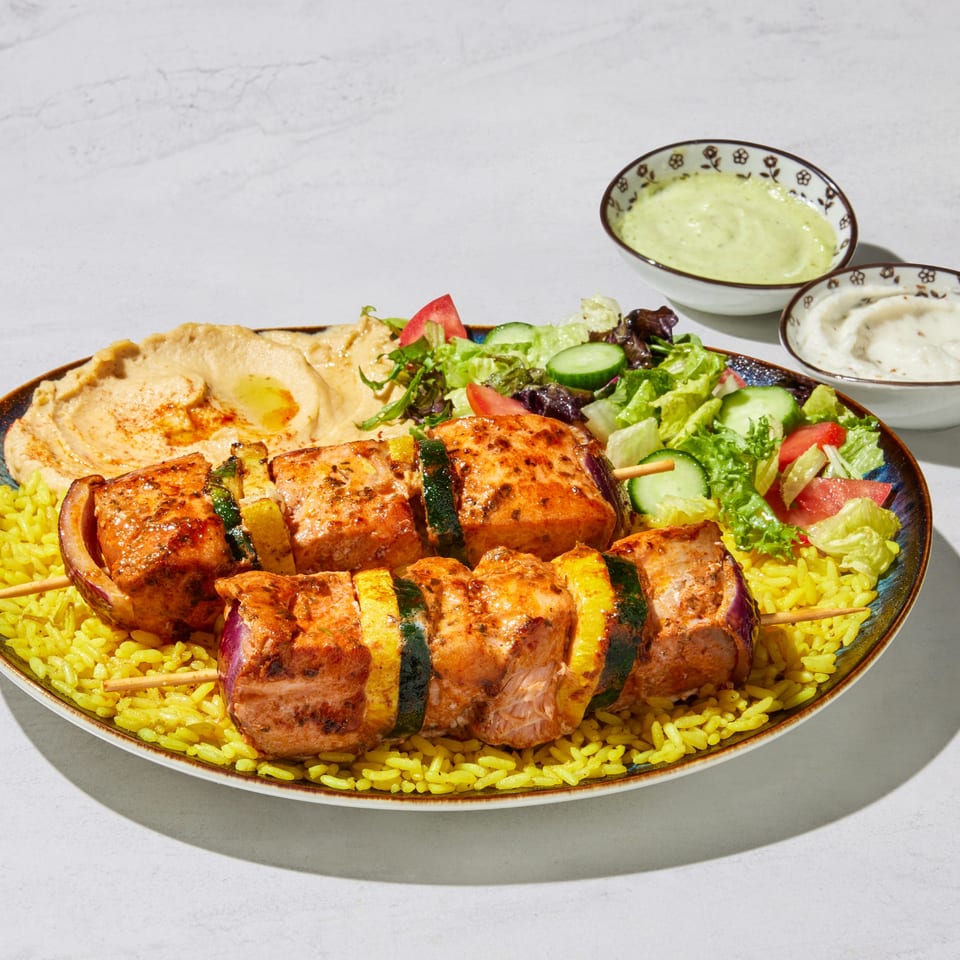Two salmon and vegetable skewers on yellow rice, with hummus, salad, and two dipping sauces.